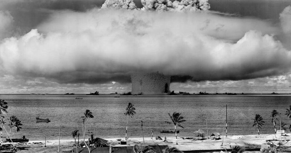 A black-and-white image of a nuclear explosion seen from a tropical beach with coconut trees.
