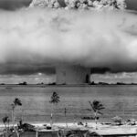 A black-and-white image of a nuclear explosion seen from a tropical beach with coconut trees.