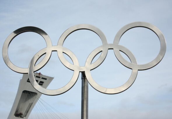 The iconic Olympic rings in front of a tall structure on a clear day.