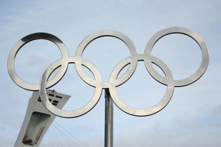 The iconic Olympic rings in front of a tall structure on a clear day.